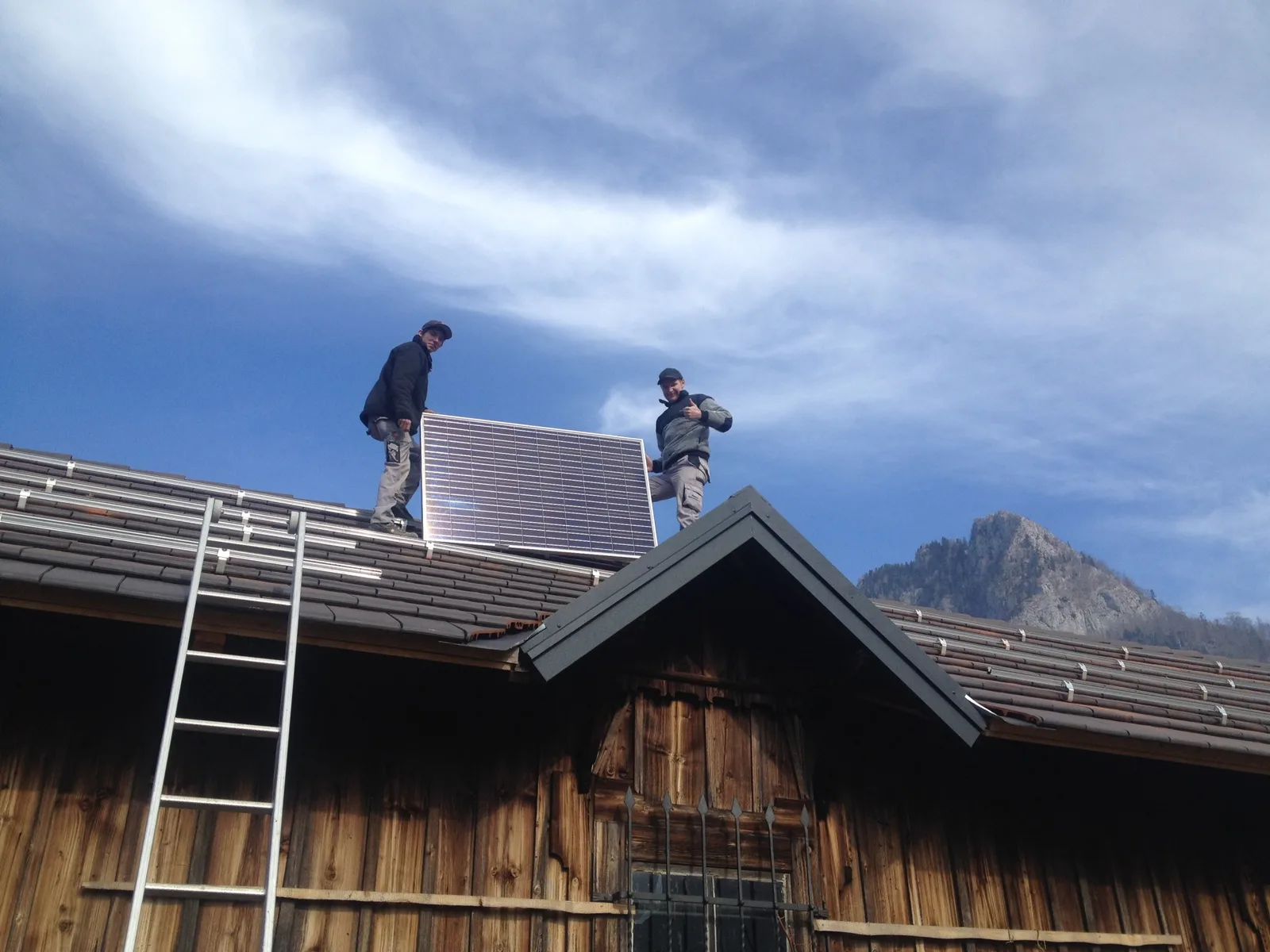 Zwei Monteure tragen ein Solarmodul über das Dach eines Holzstadels