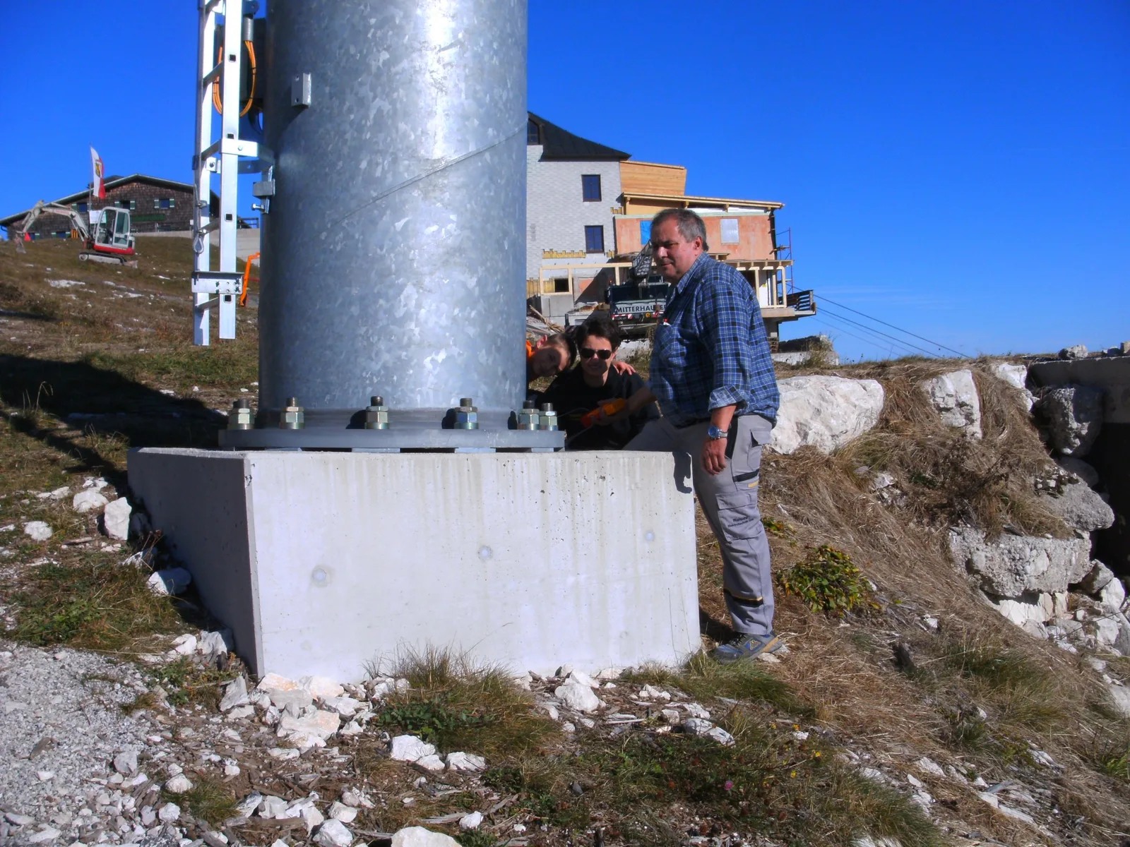 Drei Personen an einem Betonsockel unter einer verzinkten Liftstütze im Gebirge