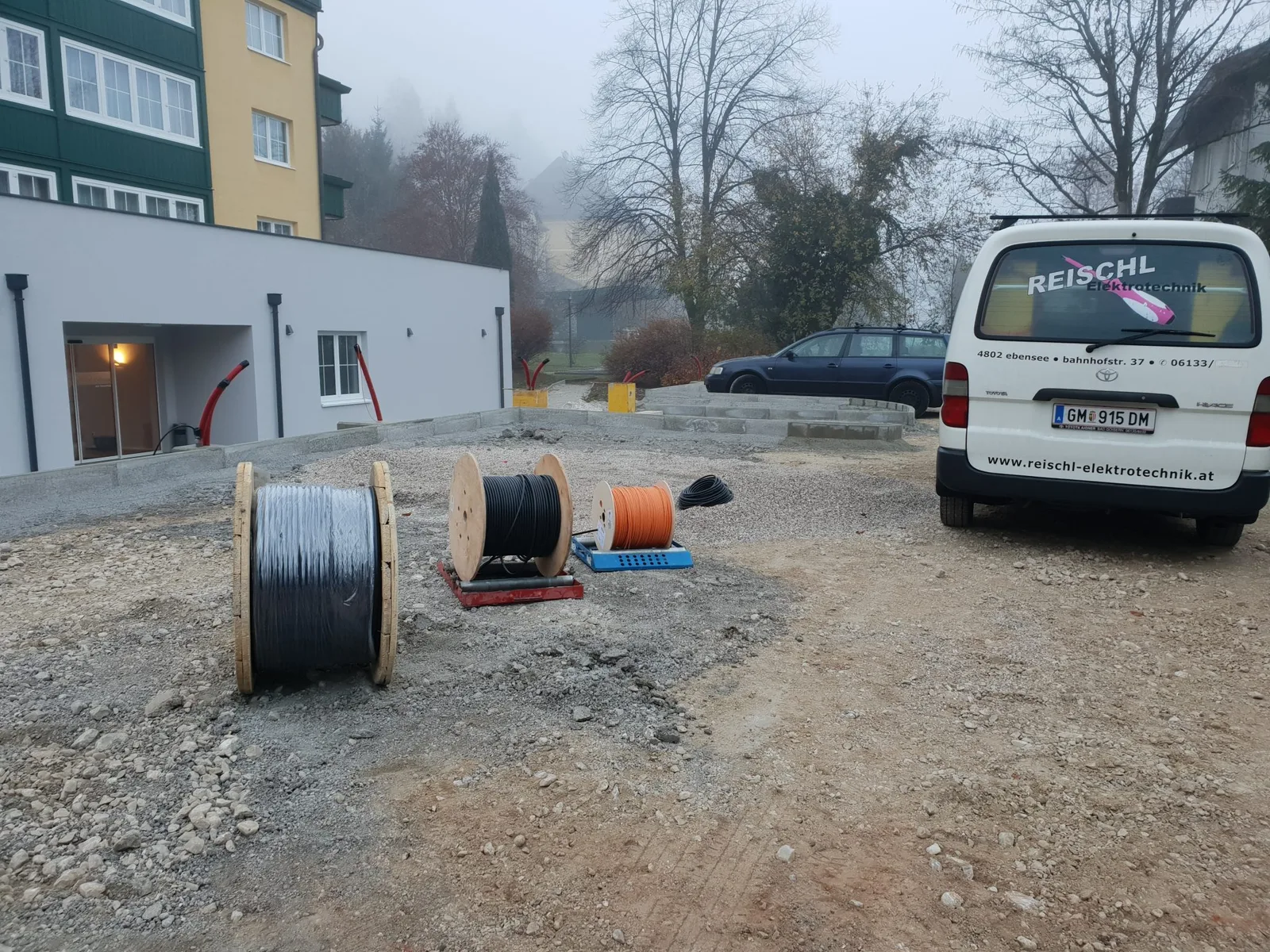 Kabeltrommeln auf einer Baustelle vor einem Gebäude, rechts ein weißer Firmenbus mit Aufschrift Reischl Elektrotechnik