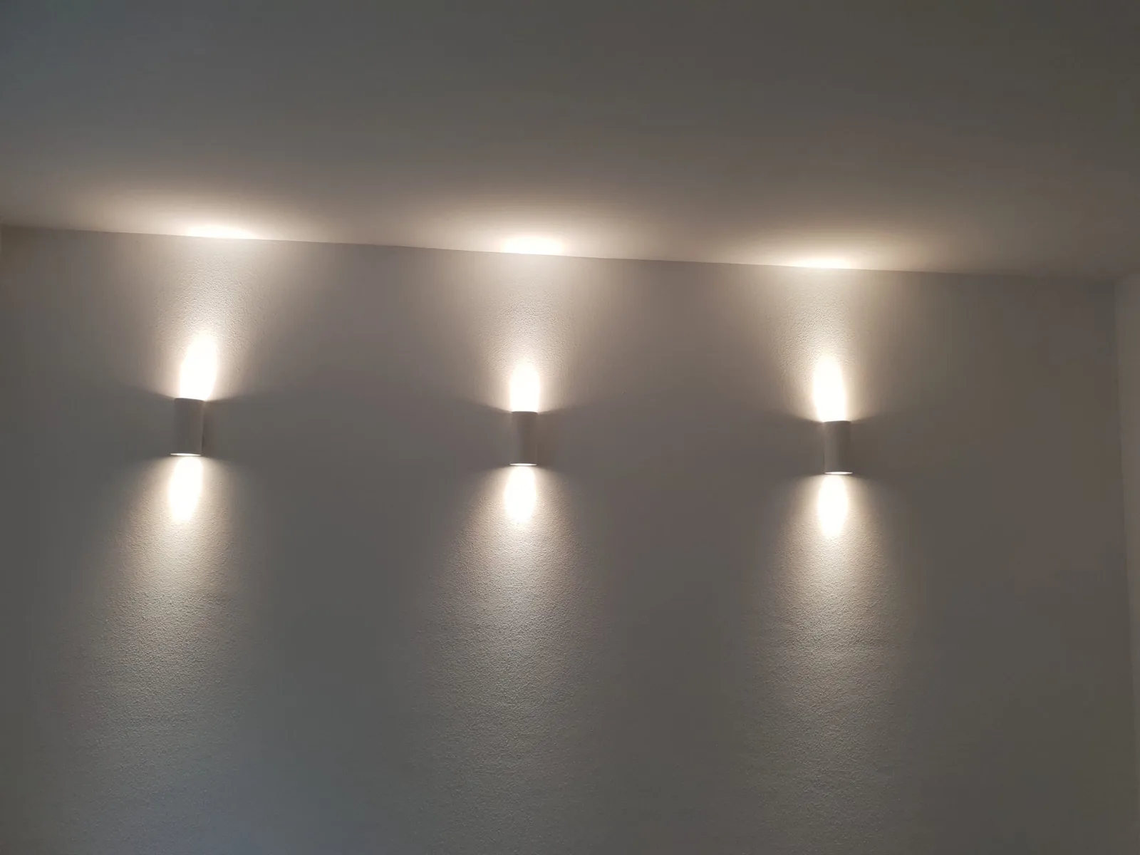 Drei Wandleuchten an einer grauen Wand, die Licht nach oben und unten werfen